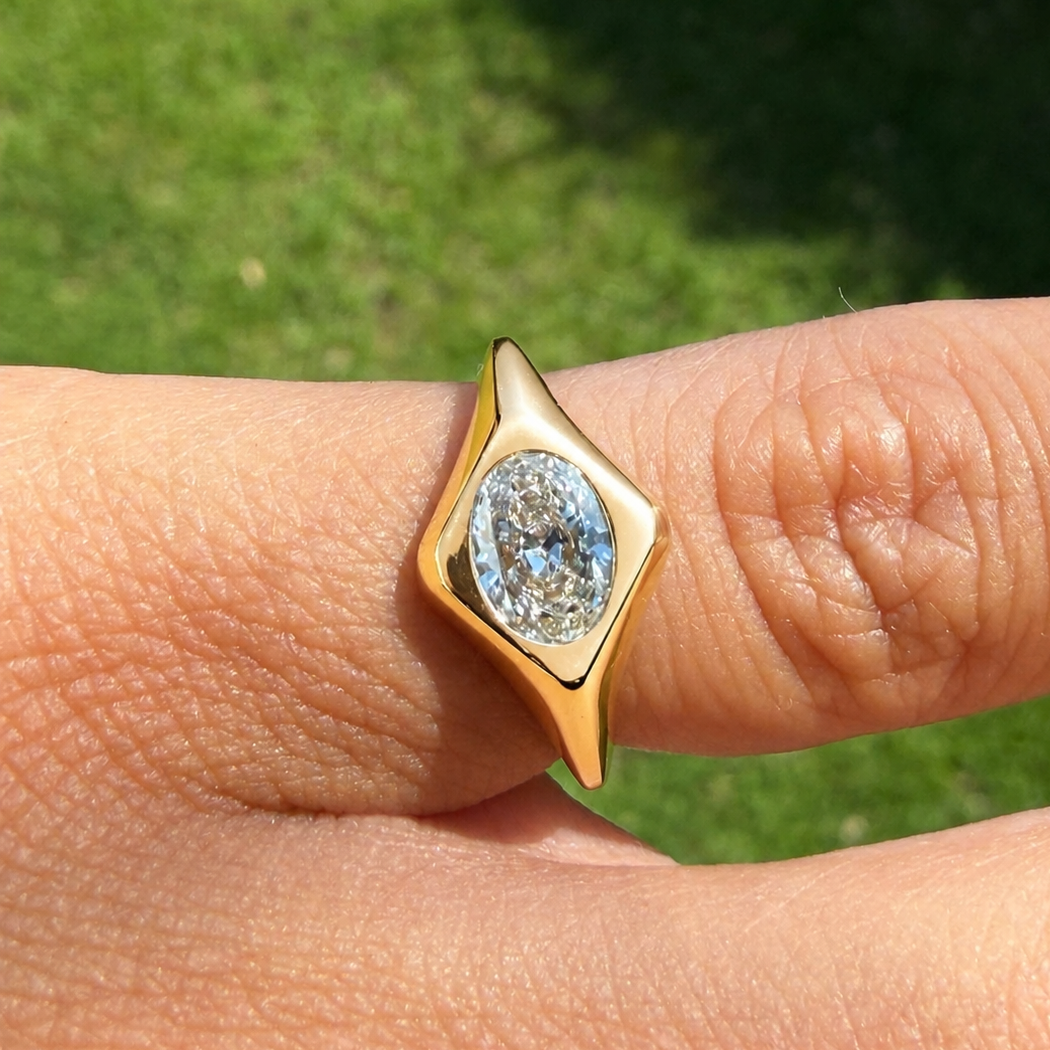 A yellow gold ring with an oval cut white gemstone set in a diamond-shaped bezel, shown worn on a finger with a blurred green grass background.