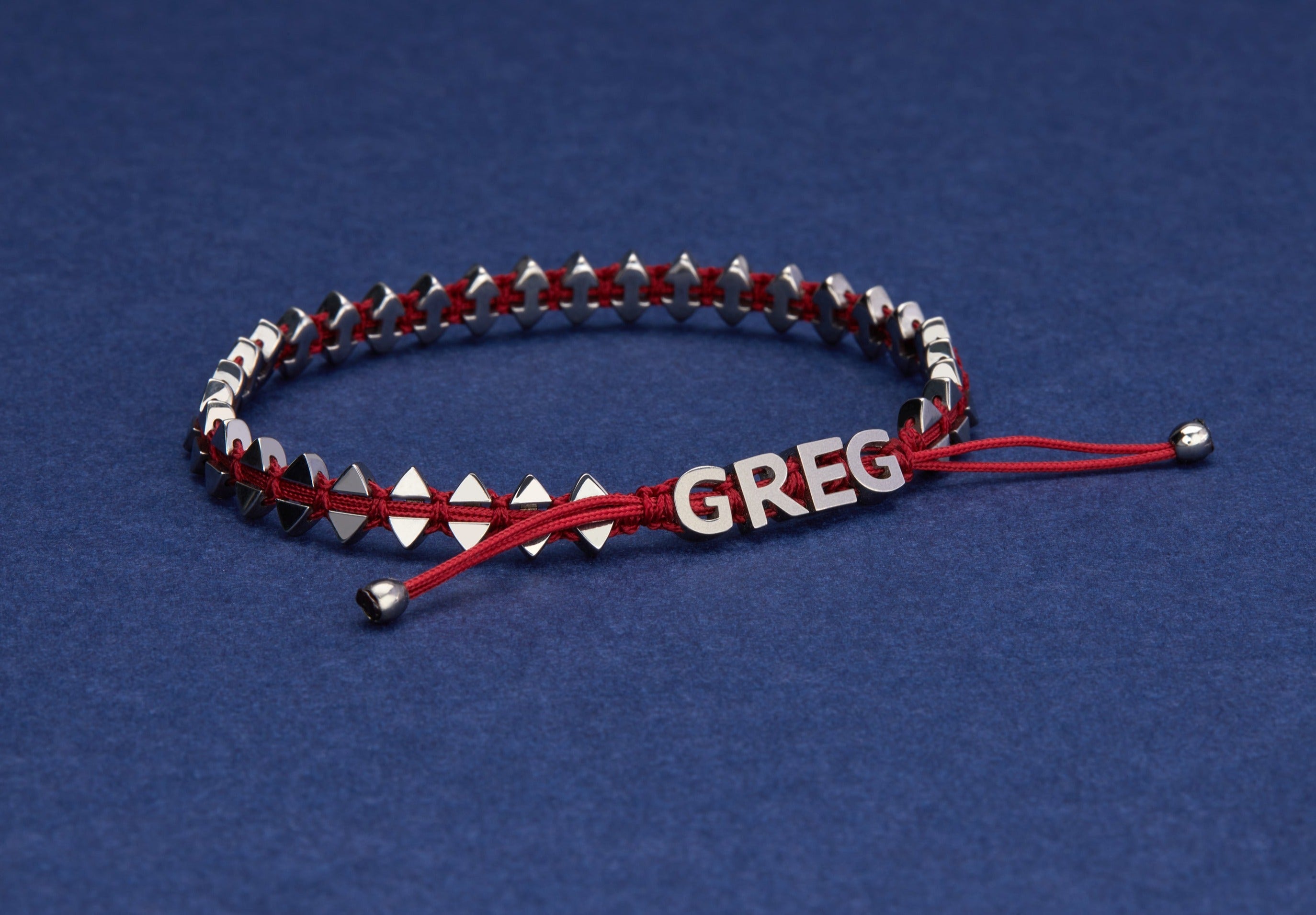 Personalised Anchor Bracelet in Carmine Red illustrating the name Greg portrayed on a blue background.