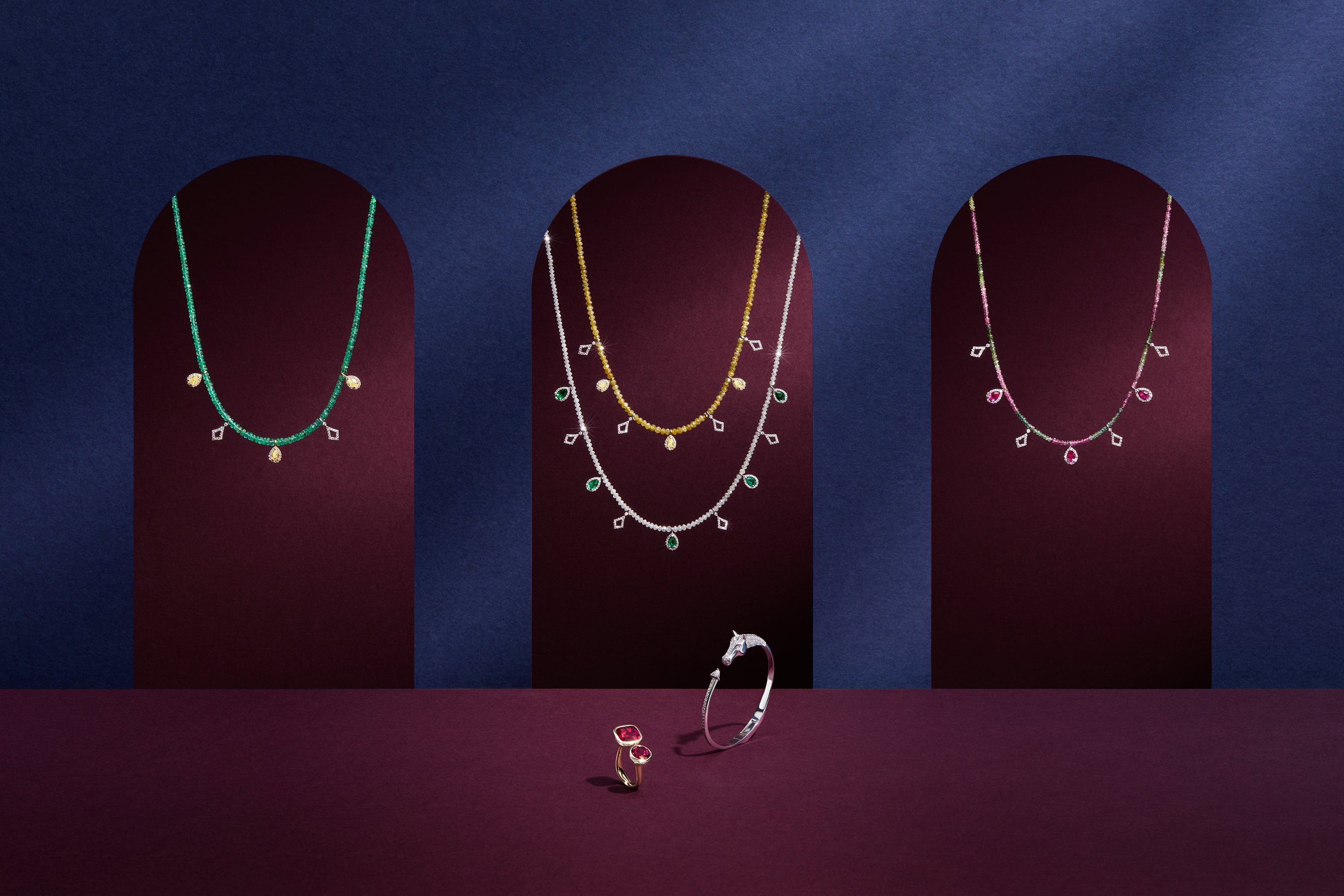 Three necklaces on dark stands against a blue background with a ring in front.