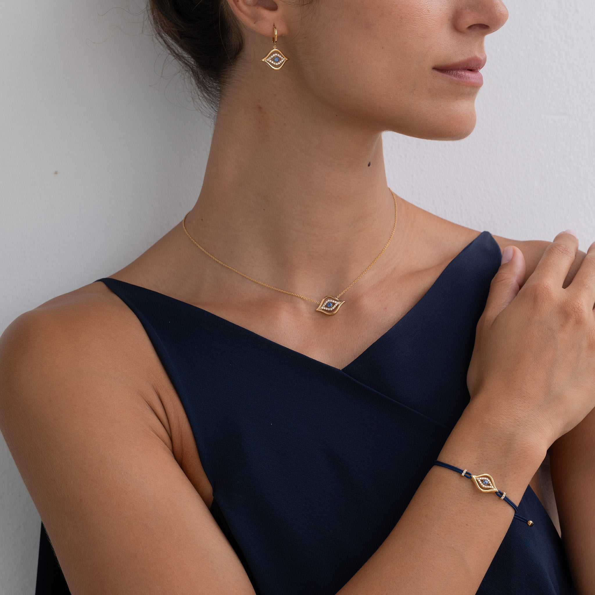 Model Wearing Evil Eye Pendant, earrings and bracelet: A model gracefully adorns the Yellow Gold Evil Eye collection with Diamonds and a round brilliant sapphire, radiating elegance and charm.
