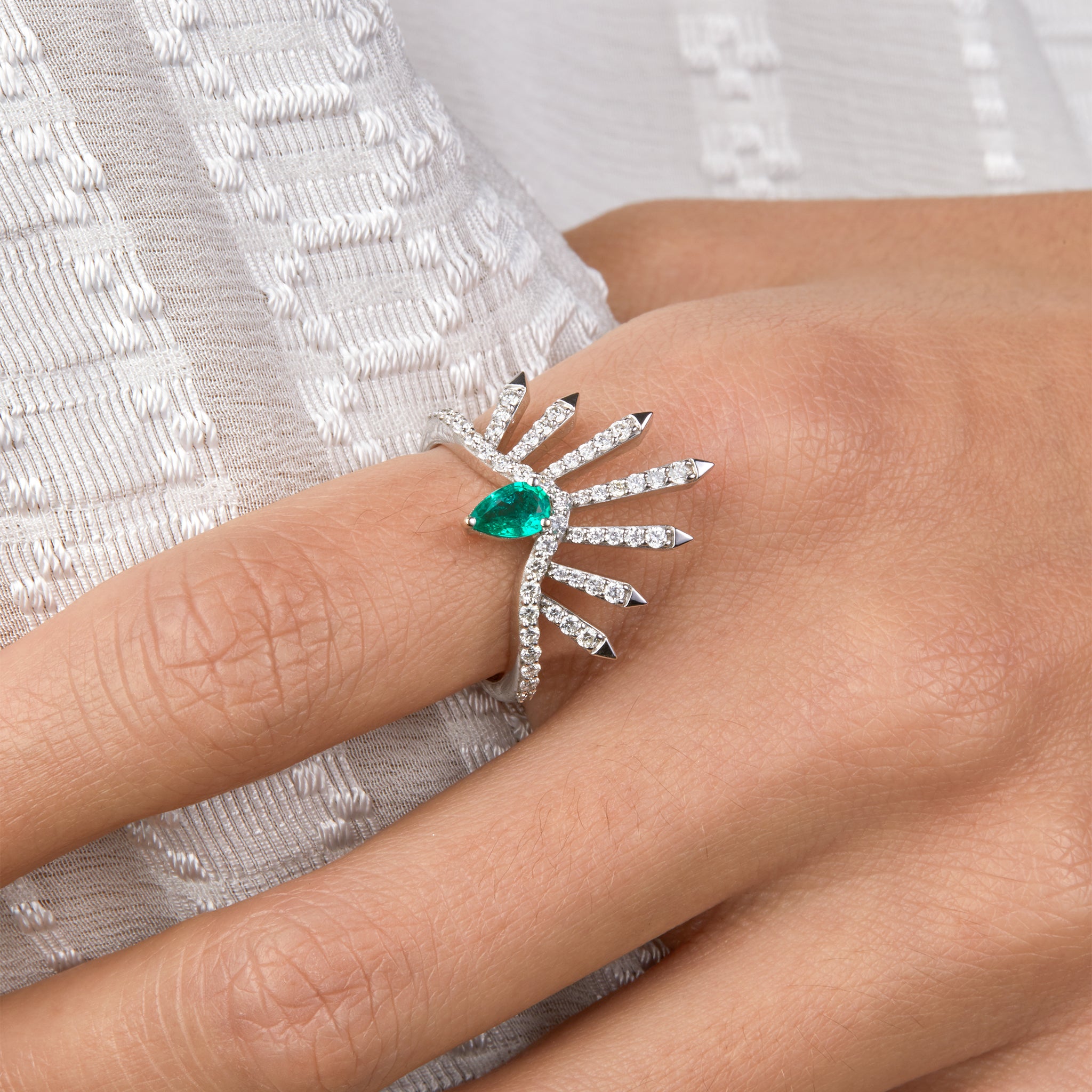 Diamond white gold ring with a pear shaped emerald portrayed facing each other on hand model wearing a denim jumpsuit.