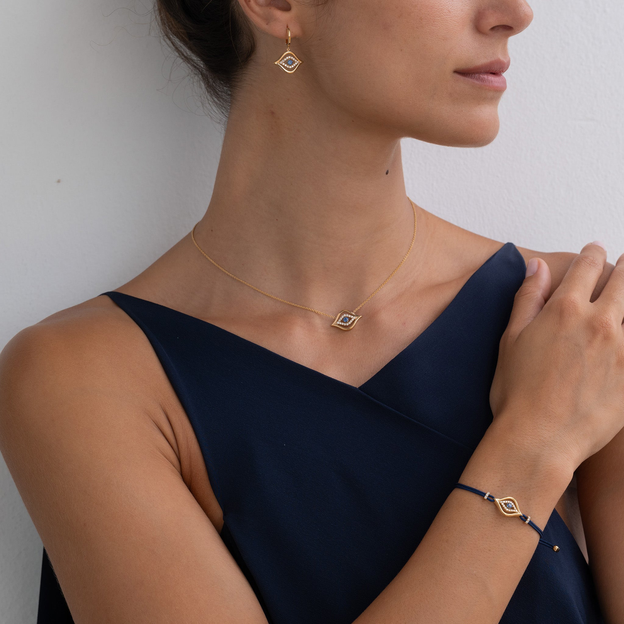 Model Wearing Evil Eye Pendant, earrings and bracelet: A model gracefully adorns the Yellow Gold Evil Eye collection with Diamonds and a round brilliant sapphire, radiating elegance and charm. 