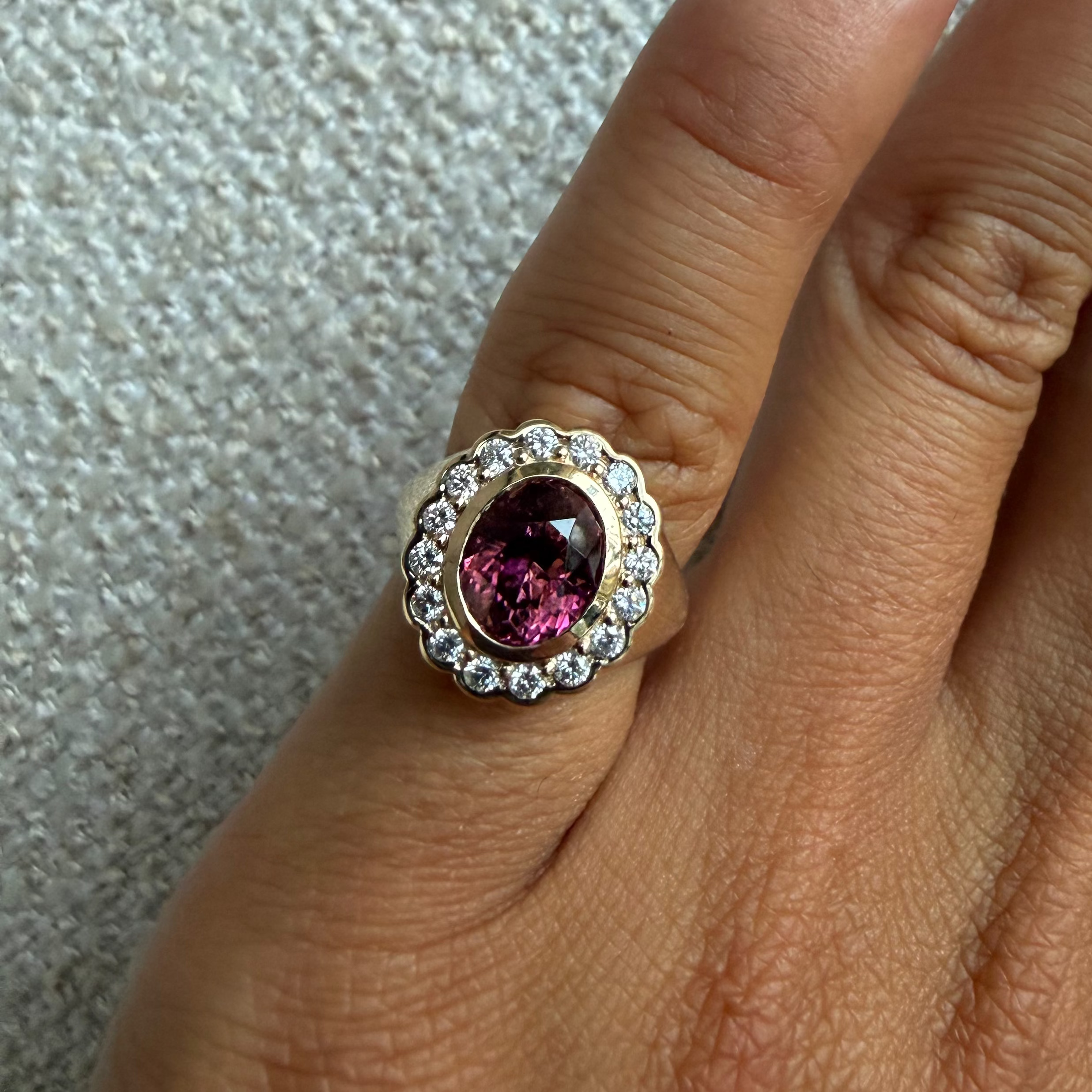 A close-up of a ring worn on a finger, featuring a scalloped yellow gold halo with small clear stones and a central oval pink gemstone.