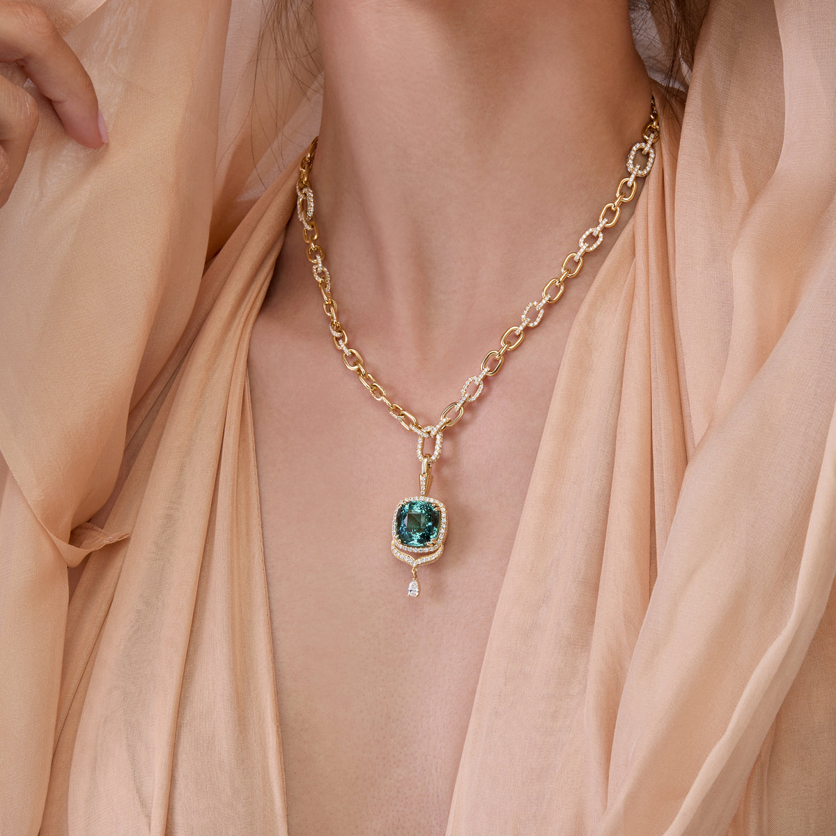 A close-up of a model wearing a gold chain necklace with a tourmaline pendant, draped in soft peach-colored fabric.