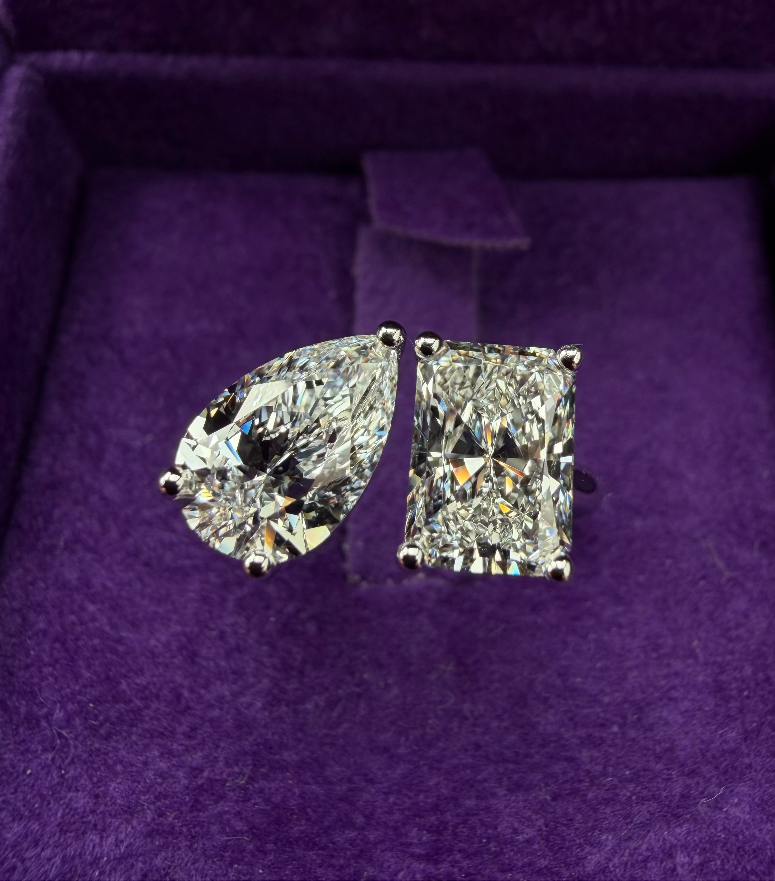 A pair of diamond rings displayed inside a purple velvet jewelry box. One ring features a pear-cut diamond, and the other features a radiant diamond, both set in white gold together.